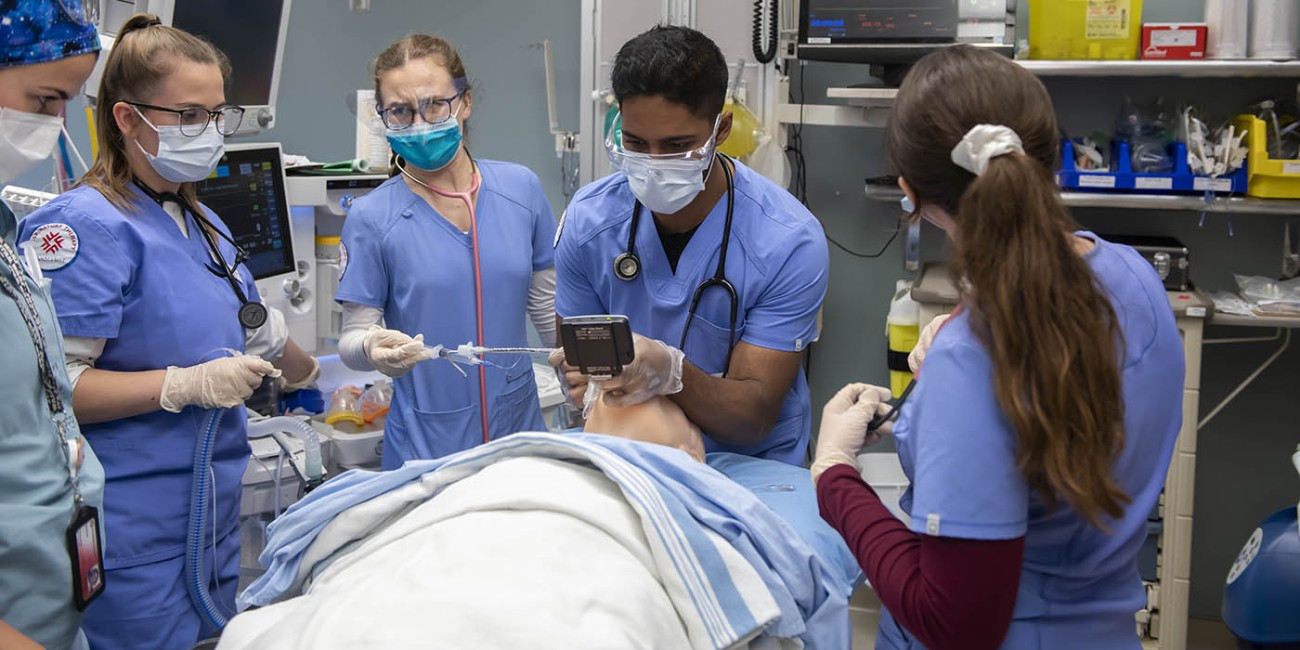 In the School of Health Sciences at Fanshawe College's London Campus, a group wearing scrubs, masks, and gloves practices intubation on a mannequin lying on a bed with blue and white striped linens. Medical equipment and monitors are visible in the background.
