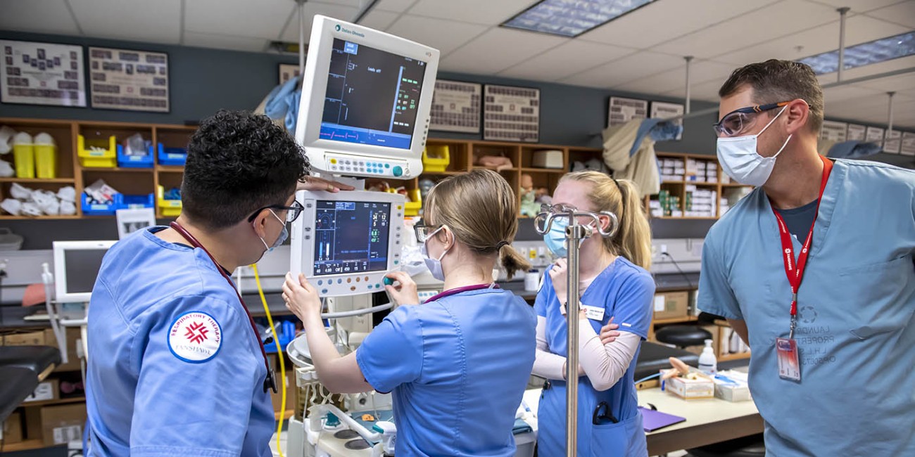 In the School of Health Sciences at Fanshawe College's London Campus, four people in blue scrubs and face masks are gathered around medical equipment, including a ventilator and monitors displaying medical data, with a mannequin lying on a table in the foreground.