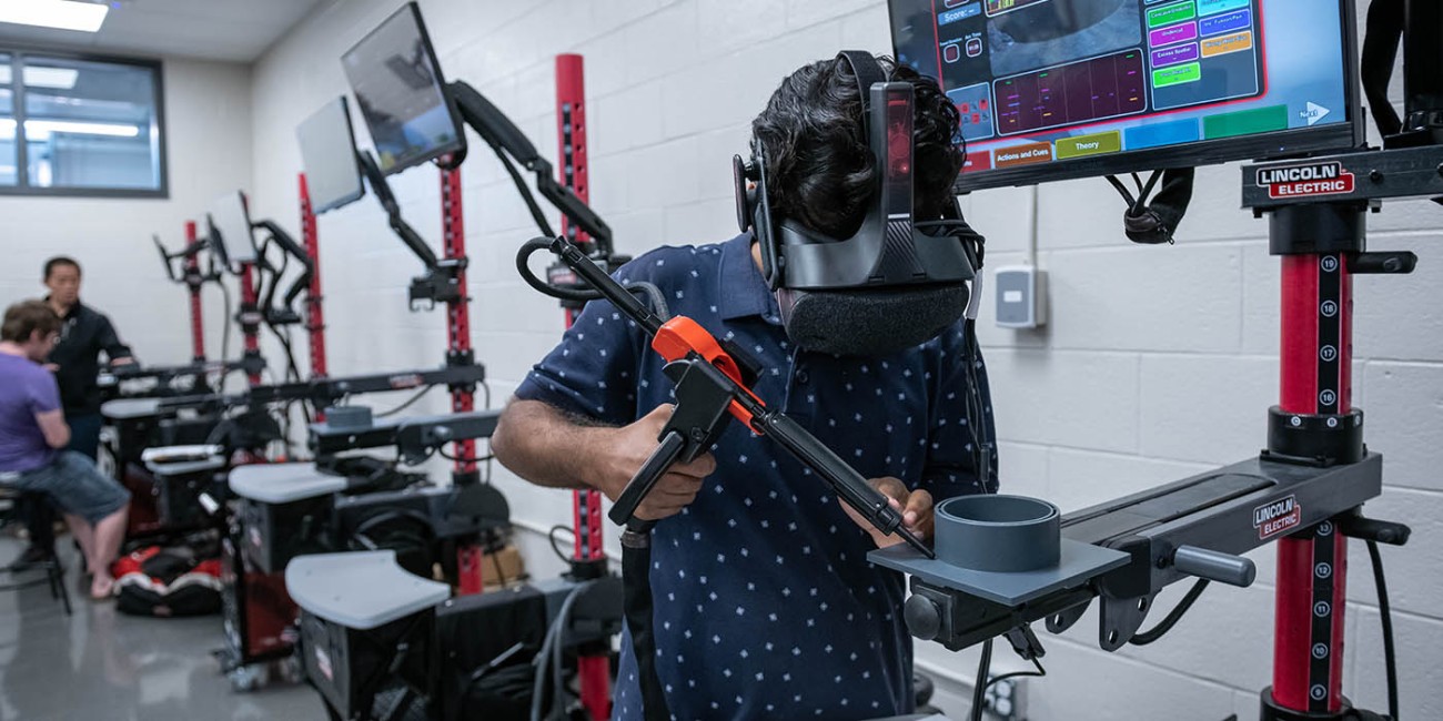 In a Fanshawe College lab, a person wearing a virtual reality headset holds a welding simulator tool, standing in front of a Lincoln Electric welding simulator with a monitor displaying welding data. Other welding simulator stations are visible in the background.