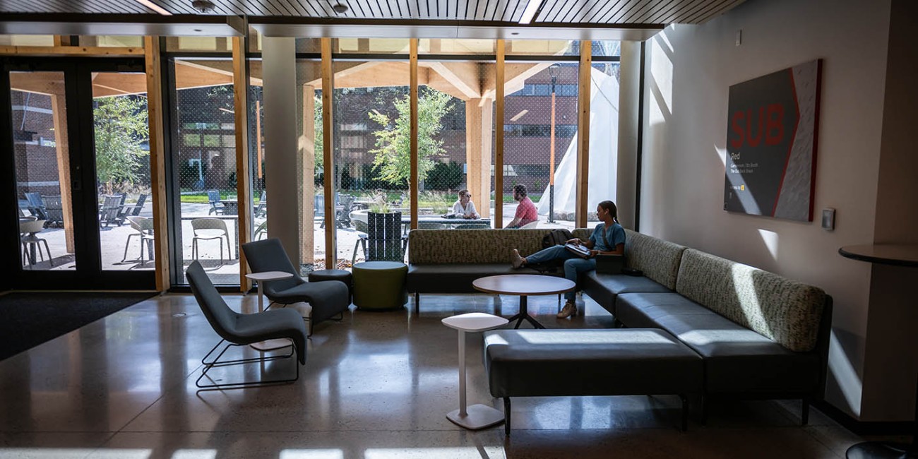 Natural light streams into the Student Union Building at Fanshawe College as a student sits lounged on an U shaped couch, next to floor to ceiling windows looking out onto a courtyard. 
