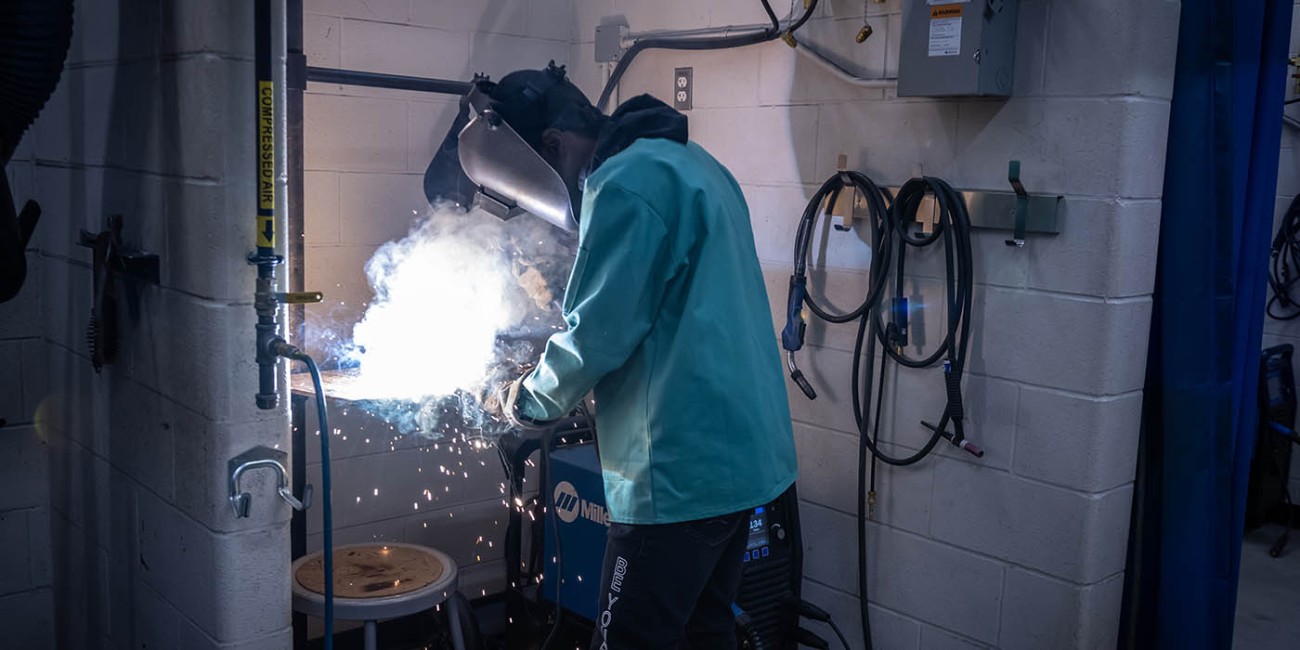 In a Fanshawe College welding lab, a person in protective gear welds metal, creating bright sparks and smoke. Welding equipment, compressed air lines, and electrical outlets are visible on the cinder block walls.