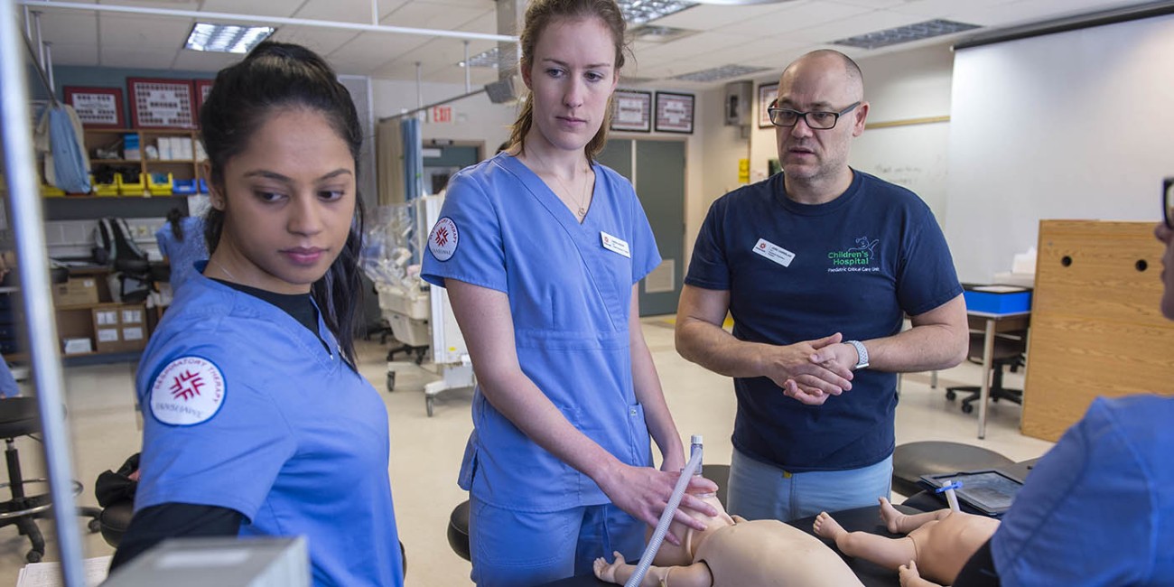 At the School of Health Sciences of Fanshawe College at the London Campus, three people stand around a black table with two baby mannequins, as one person in blue scrubs holds a mask connected to a tube over the face of one of the mannequins.