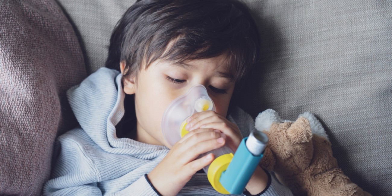 A young child sits amongst the cushions of a couch, holding an MDI inhaler with a spacer as a supportive stuff animals lays close by.