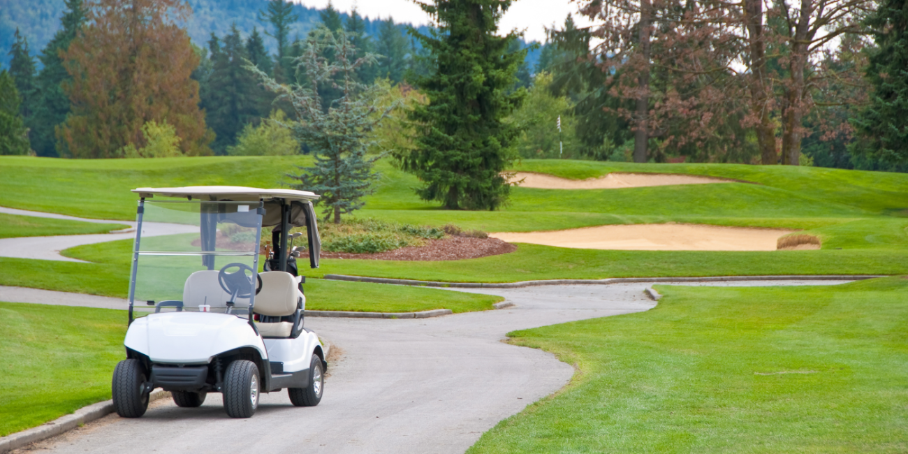 An electric golf cart travels down a pathway on a golf course between two greens, with trees and sand bunkers in the distance