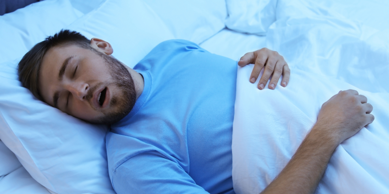 A man in a blue shirt sleeps with his mouth agape on a white pillow covered by a white duvet