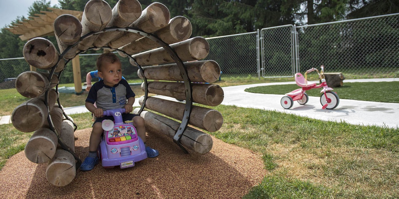 An individual sits on a purple toy car under a log tunnel at the Early Childhood Education program at the Simcoe/Norfolk Regional Campus, with a pink tricycle visible in the background.