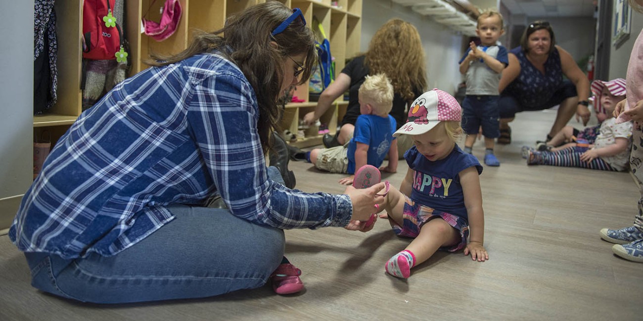 Inside the Simcoe/Norfolk Regional Campus, Early Childhood Education program, an individual wearing a blue plaid shirt and jeans kneels on the floor, helping a child put on a pink shoe. Other children and adults are nearby, with cubbies 