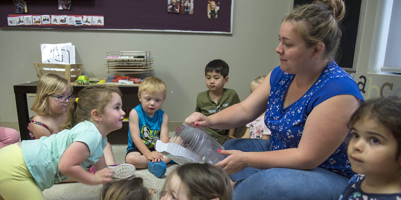In a classroom at the Simcoe/Norfolk Regional Campus, an educator sits cross-legged on the floor, holding a clear plastic container, surrounded by children who are also sitting on the floor. The children are looking at the container and each other. Behind them is a low table with toys and learning materials, and a bulletin board with pictures and visual aids. The letters "abc" are visible on the wall. This is likely an activity for the Early Childhood Education program.