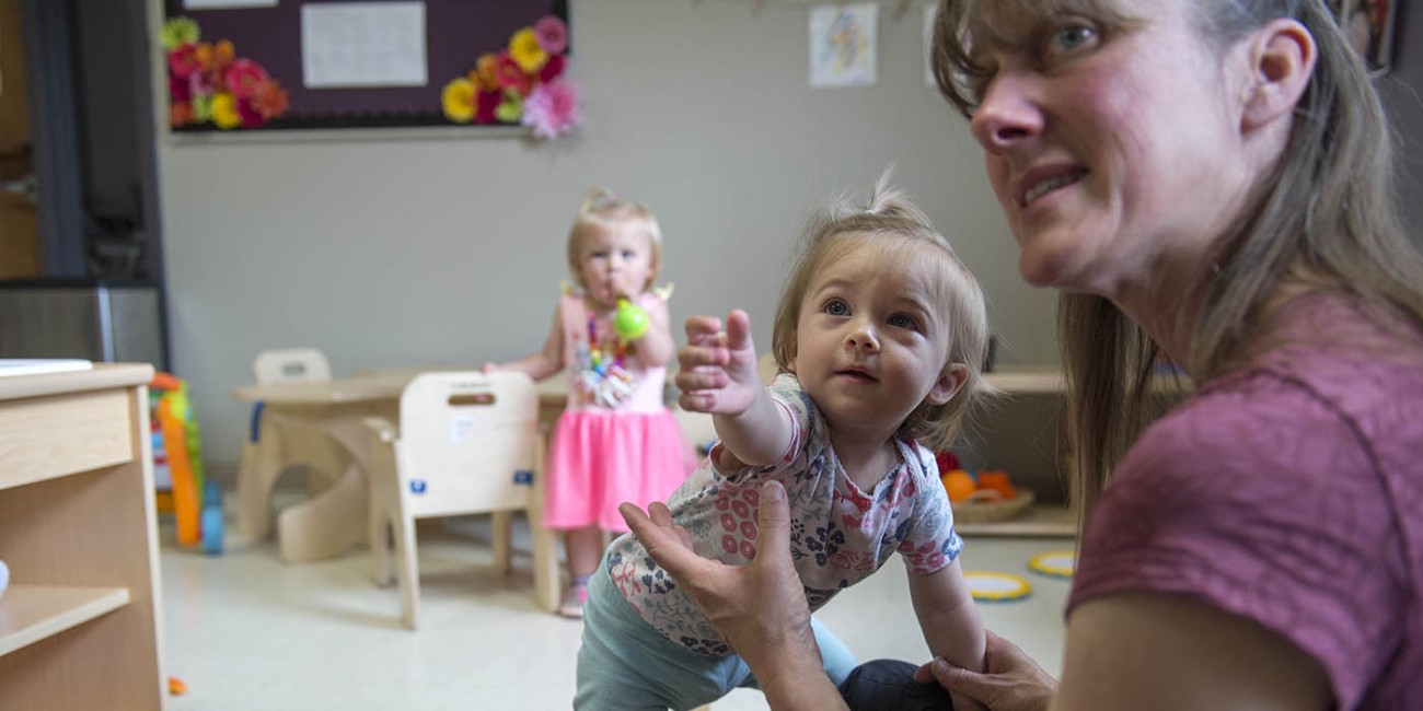 At the Simcoe/Norfolk Regional Campus, a person wearing a maroon shirt is helping a toddler wearing a floral shirt and blue pants stand, with their arm outstretched, in a room with a basket of wooden blocks, a small table and chairs, and another toddler in a pink dress.