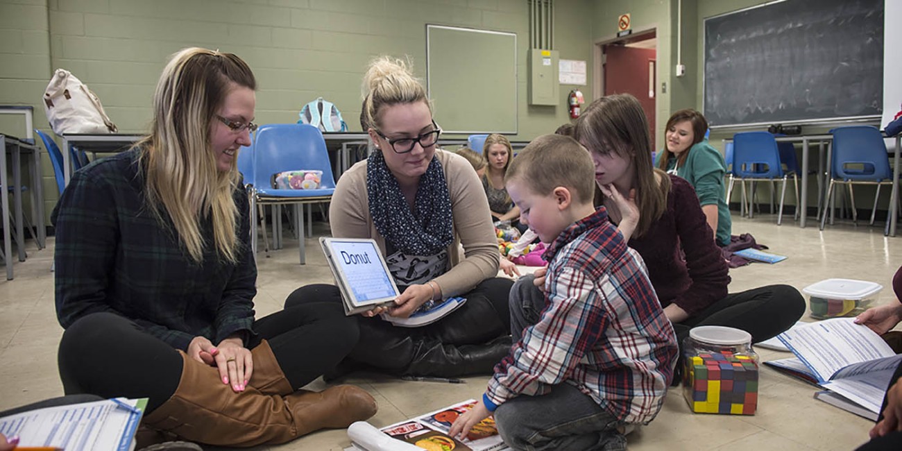 In a classroom at Fanshawe College's School of Community Studies, a group sits on the floor, looking at books and tablets, with a child pointing at a picture in a book.