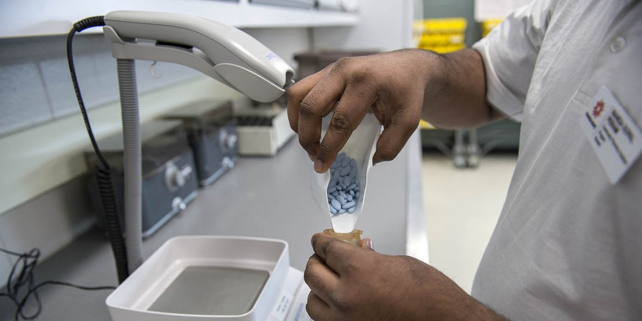At the School of Health Sciences of Fanshawe College at the London Campus, a person wearing a white shirt and an ID badge pours blue pills from a small plastic bag into a small amber bottle, while standing at a counter with a scale, a white tray, and a white bottle with a blue symbol on the lid.