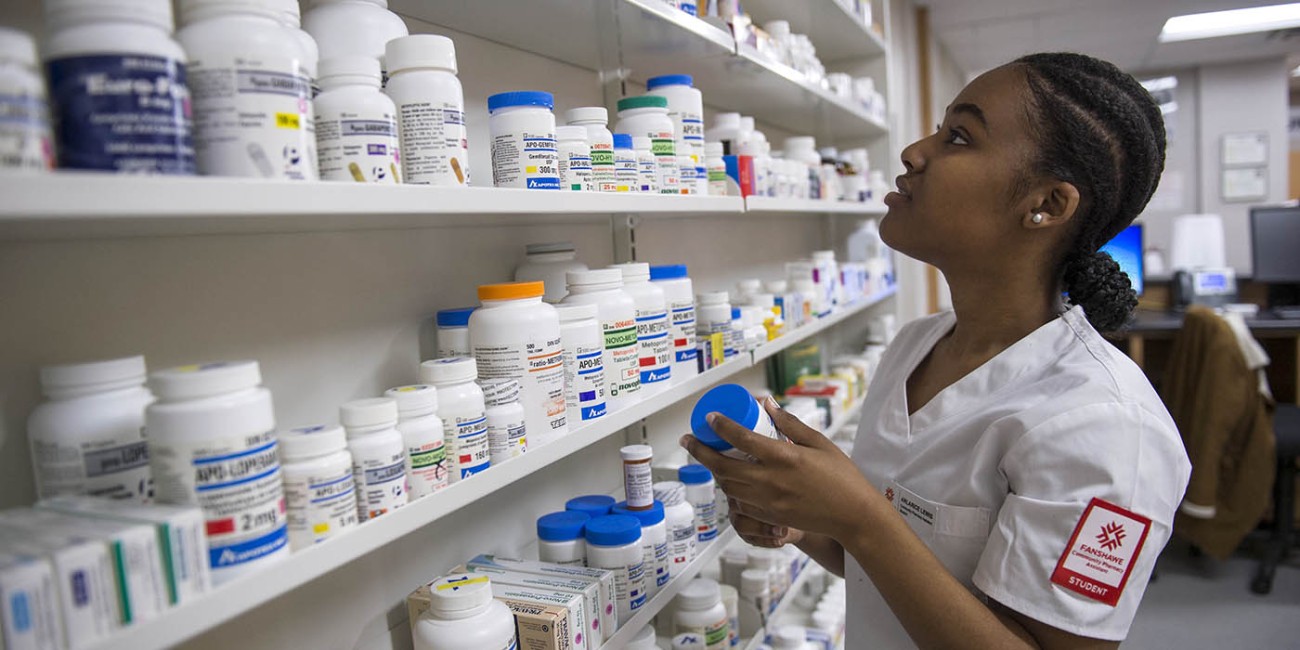 Inside the School of Health Sciences at Fanshawe College's London Campus, a person wearing a white uniform with a "Fanshawe Community Pharmacy Assistant STUDENT" patch on the sleeve reaches for a bottle on a shelf stocked with various medications.