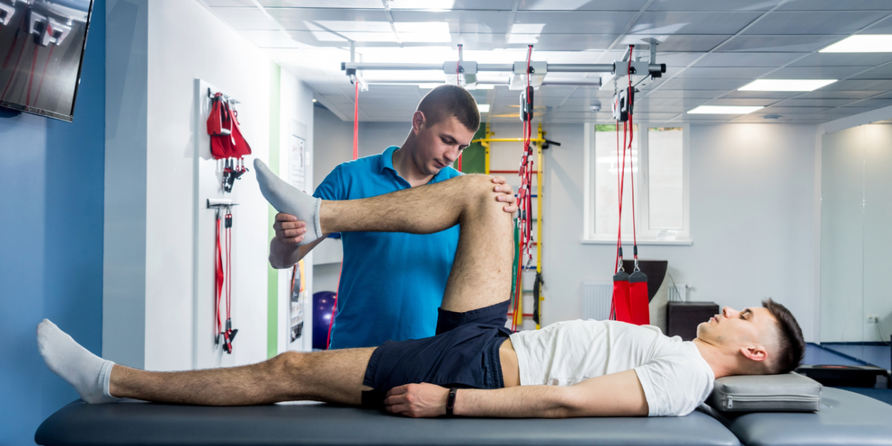 A physiotherapy practitioner works with a patient on his hip motion inside a rehab facility with various equipment in the background. 