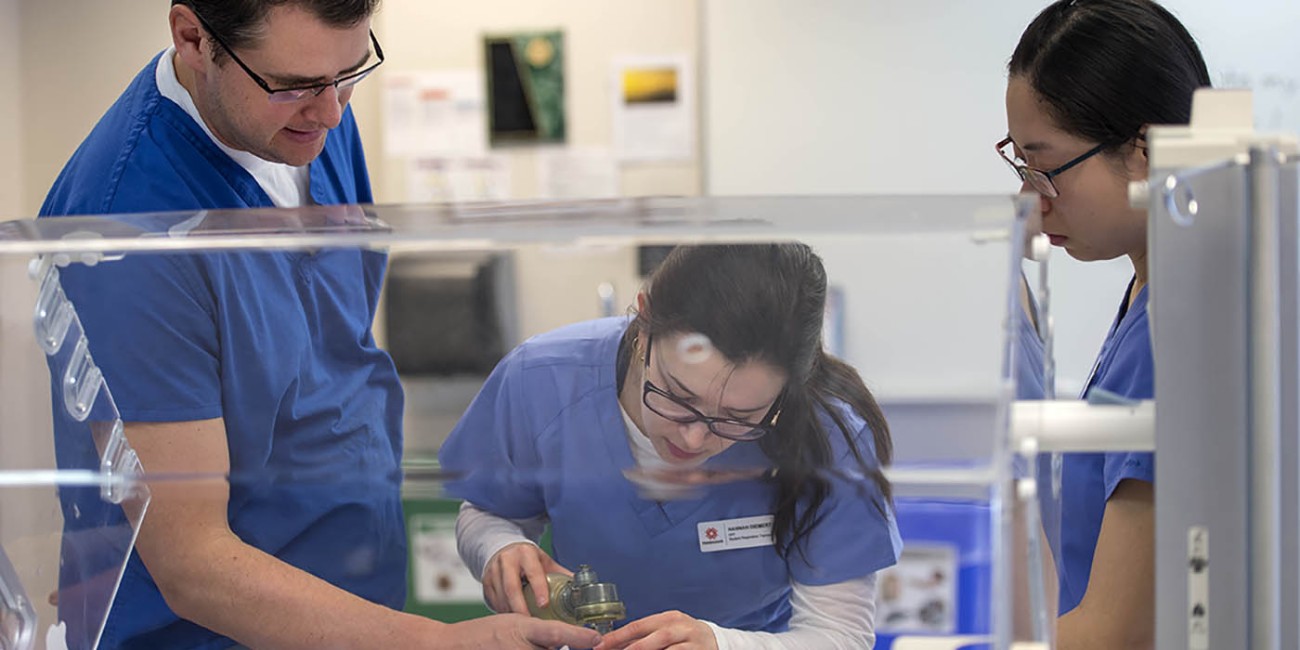 At the School of Health Sciences of Fanshawe College at the London Campus, three people in blue scrubs are gathered around a clear incubator, with one person holding a medical device over a covered form inside