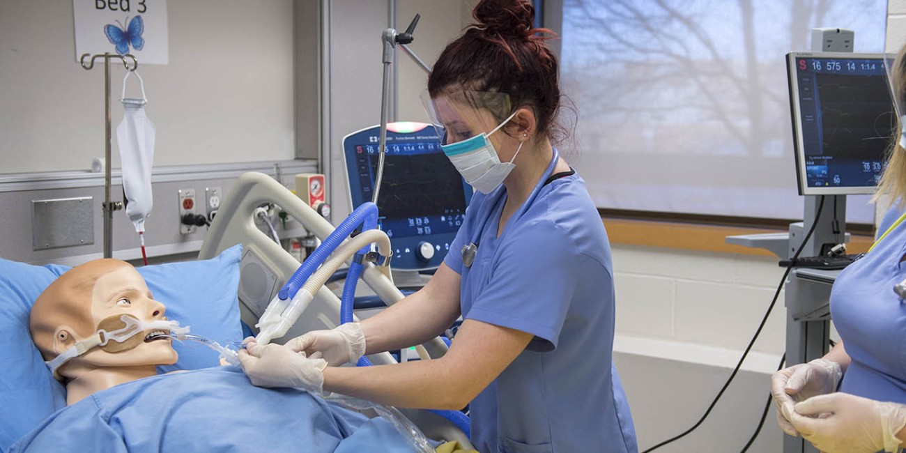 At the School of Health Sciences of Fanshawe College at the London Campus, a person wearing a face shield, mask, gloves, and blue scrubs is working with a medical mannequin in a hospital bed, connected to a ventilator and a monitor displaying medical data.