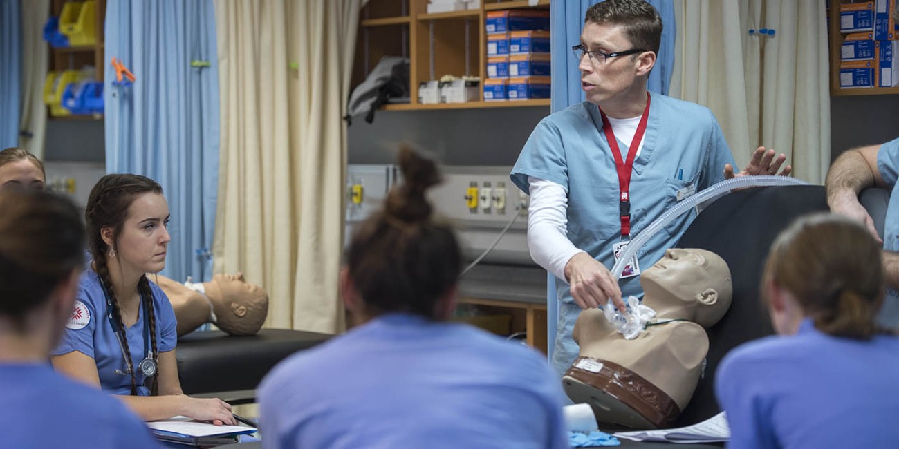 In a classroom at Fanshawe College's School of Health Sciences at the London Campus, a person in scrubs demonstrates a medical procedure on a mannequin to a group of people, also in scrubs. The mannequin is lying on a black surface, and the demonstrator is holding a clear tube connected to the mannequin's neck. The classroom has blue and beige curtains, and shelves with medical supplies.