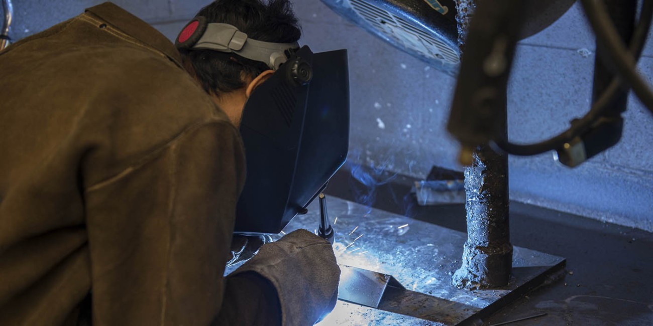 At Fanshawe College, a person wearing a welding mask, gloves, and jacket is welding metal on a workbench.