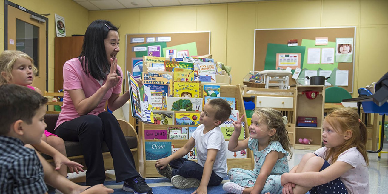 In a classroom at the Goddard School, a group of children sit on a blue and white striped rug, listening to someone reading a book. Behind them is a bookshelf filled with colorful children's books, and bulletin boards with various papers and pictures.