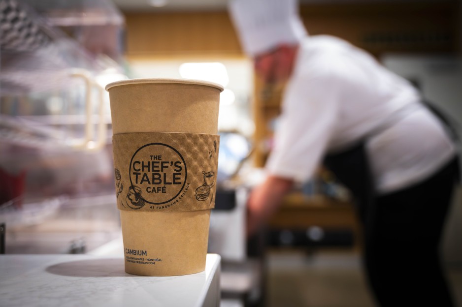 A paper cup with a sleeve that reads "The Chef's Table Cafe at Fanshawe College" sits on a counter at the London Downtown Campus, with a chef in the background.