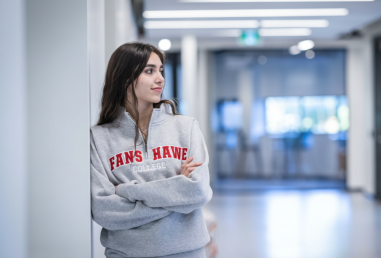 A person wearing a grey sweater with red lettering leans against a white wall in a hallway.