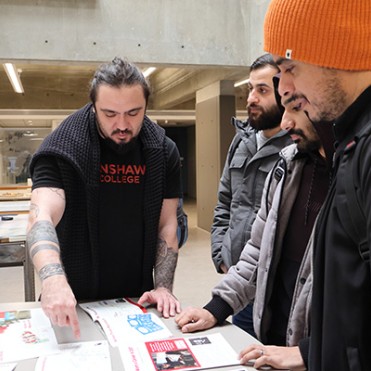 A group of four men gather around a table, focused intently on documents. One man with tattoos gestures towards the papers, explaining something. The setting is a modern, well-lit space, conveying a collaborative and educational atmosphere.