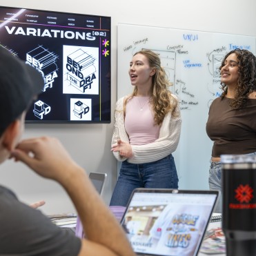 A group of individuals is gathered in a room, facing a large screen displaying "COLOUR VARIATIONS" and graphic designs, including a "BEYOND THE DRAFT" logo. To the right, a whiteboard is covered with UX/UI notes and diagrams. Two people stand near the screens, one speaking with hands gesturing, the other smiling. In the foreground, the back of a person's head and shoulder are visible, along with open laptops and a black travel mug on a table.