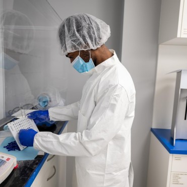 At the School of Health Sciences of Fanshawe College at the London Campus, a person wearing a hair net, face mask, blue gloves, and a white lab coat works inside a clear enclosure, holding a white tray with holes and blue capsules.