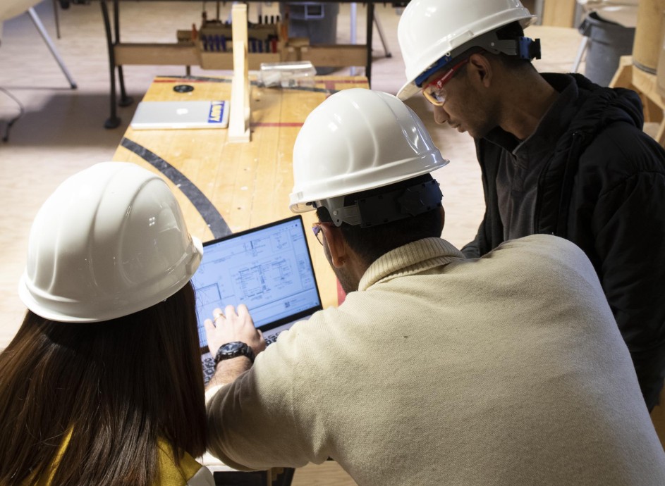 Two students with Construction Project Management instructor, reviewing plans on laptop at job site