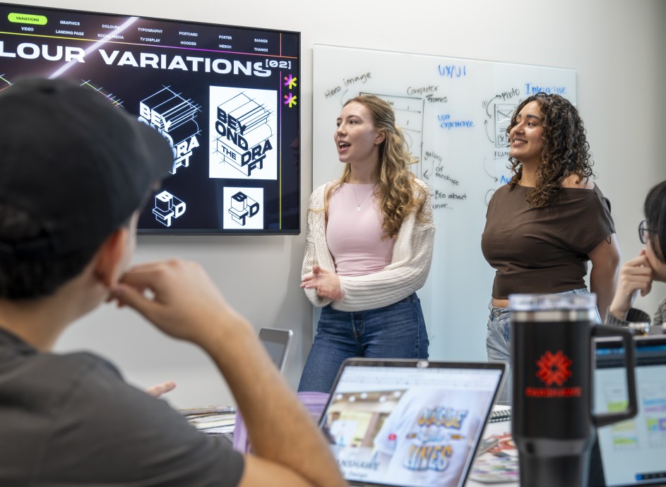 A group of individuals is gathered in a room, facing a large screen displaying "COLOUR VARIATIONS" and graphic designs, including a "BEYOND THE DRAFT" logo. To the right, a whiteboard is covered with UX/UI notes and diagrams. Two people stand near the screens, one speaking with hands gesturing, the other smiling. In the foreground, the back of a person's head and shoulder are visible, along with open laptops and a black travel mug on a table.