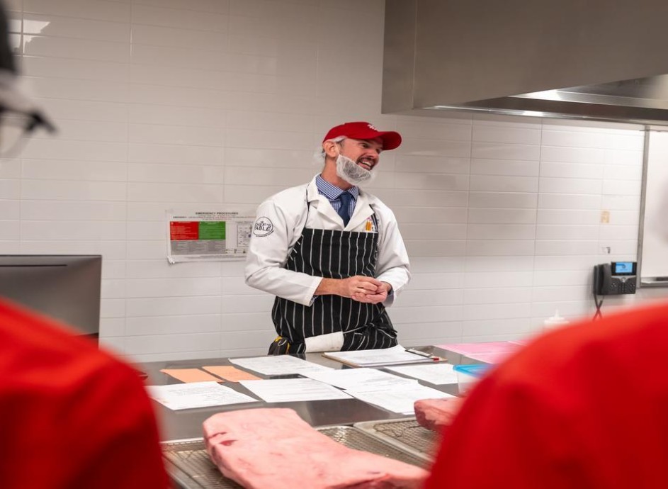 At the London Downtown Campus, in a room with white tiled walls and stainless steel counters, a person in a white lab coat, red baseball cap, and black and white striped apron stands behind a counter with papers and a large cut of meat, while two people in red jackets stand in the foreground.