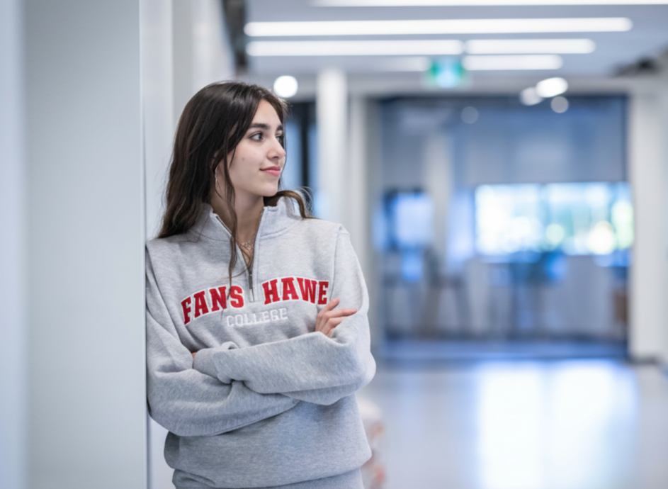 A person wearing a grey sweater with red lettering leans against a white wall in a hallway.
