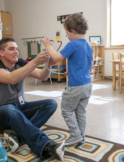 Early Childhood educator sits on the floor interacting with a toddler in a classroom