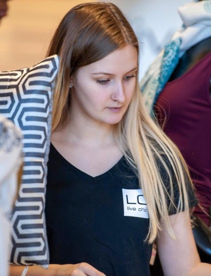 A woman with long blonde hair and a black "LOGI" shirt stands near a mannequin draped in scarves, holding a patterned pillow. She appears focused.