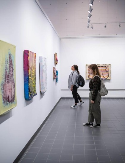 Two people in a modern art gallery view colorful, textured artworks on a white wall. The atmosphere is calm, with a focus on artistic appreciation.