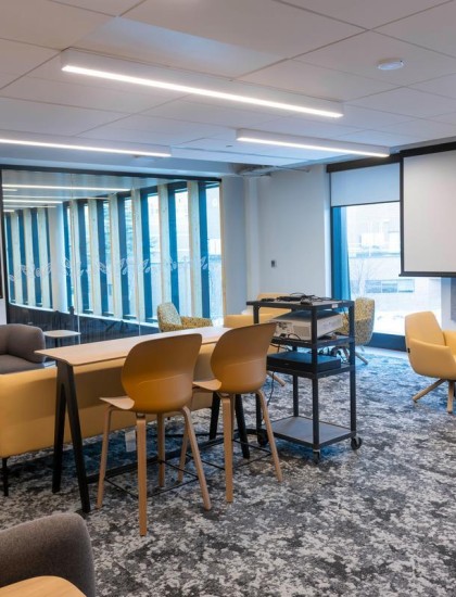 Modern office lounge with gray sofas, yellow chairs, a large screen, and floor-to-ceiling windows. The space feels open and inviting.