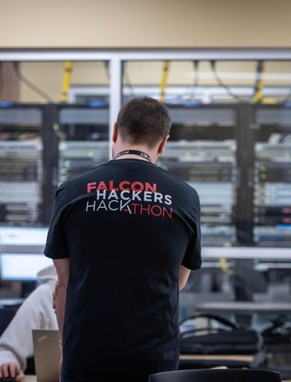 A person in a "Falcon Hackers Hackathon" shirt stands facing computer servers. Another person is nearby handling cables, creating a focused tech atmosphere.