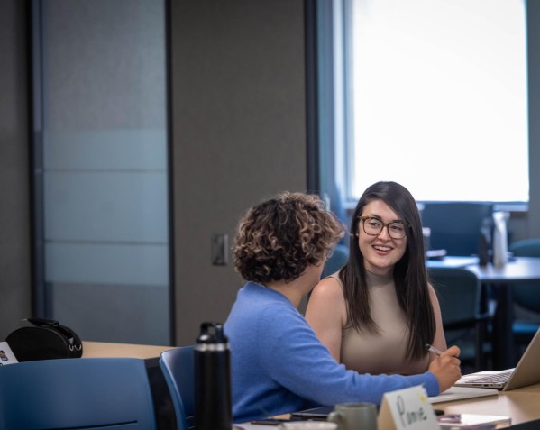 Two individuals sit at a table with laptops and notebooks, engaged in conversation.