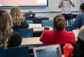In a classroom setting, an individual stands near a whiteboard covered in notes, gesturing towards a projected screen displaying text, while others sit at desks with laptops.