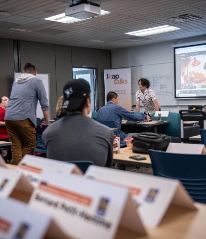 A classroom setting with tables and blue chairs, a projector screen displaying a video call, and a whiteboard with notes.
