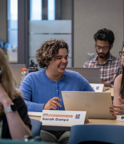 Several people sit at tables in a classroom setting, working on laptops and engaging in conversation.