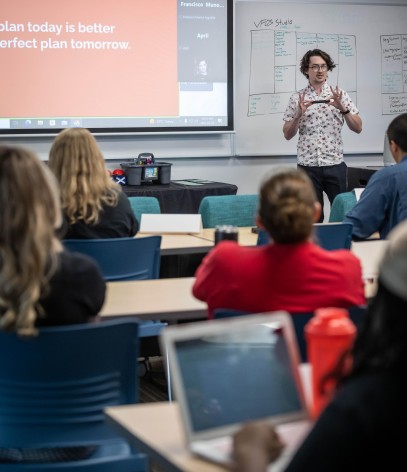 In a classroom setting, an individual stands near a whiteboard covered in notes, gesturing towards a projected screen displaying text, while others sit at desks with laptops.