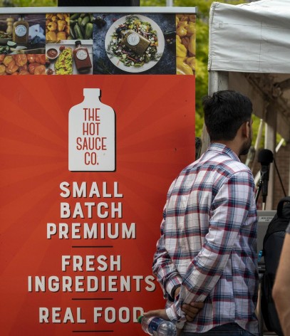 A bright orange sign advertises hot sauce with white text and a white bottle outline, with images of food and peppers above the sign.