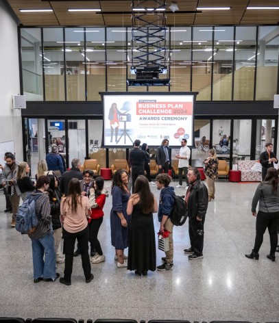 A large group of people are gathered in a spacious hall with a high ceiling and large windows, with a screen displaying information about a business plan challenge awards ceremony.