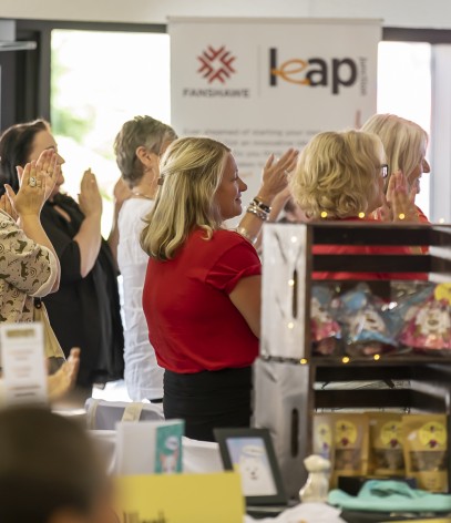 A group of people are clapping, with a banner and a display of items visible in the foreground.
