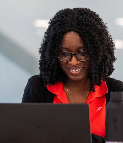 A woman with curly hair and glasses smiles while using a laptop. She wears a red shirt and sits in a modern space with yellow chairs.