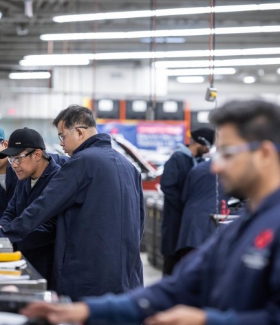 At the Centre for Applied Transportation Technology at Fanshawe College, several people in navy blue work shirts and safety glasses are gathered around a workbench with a computer, tools, and papers, while other people work on vehicles with open hoods in the background.