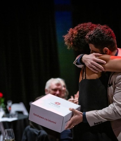 Two people embrace while one holds a Fanshawe Alumni box.