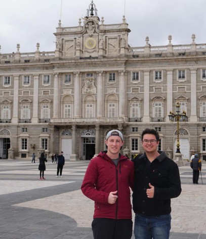 Two individuals stand in front of a large, ornate building with many windows and columns, on a grey, overcast day.