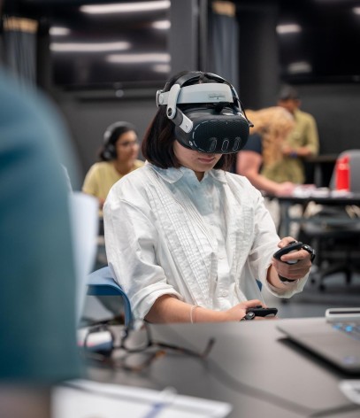 A person with short dark hair sits at a desk, wearing a white virtual reality headset and a white button-down shirt while holding handheld controllers.