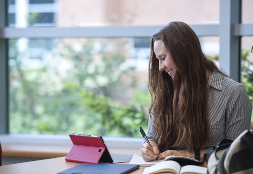 Student writing at table while looking at tablet